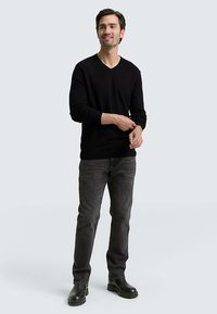 Maglione nero a V, jeans grigi e stivaletti neri. Modello in piedi, sorridente, con le mani leggermente sollevate, su uno sfondo grigio chiaro.