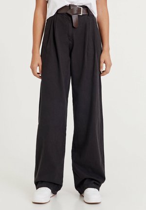 Personne portant un pantalon noir large avec des plis, une ceinture marron, des baskets blanches et un t-shirt blanc rentré, debout devant un arrière-plan uni.