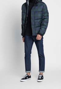 Person som bär en grön rutigt puffjacka, svart polotröja, blå jeans och svarta sneakers, stående mot en ljus bakgrund.