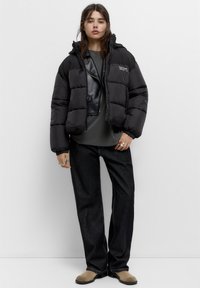 Giacca bomber nera con design corto e cappuccio, indossata sopra una maglietta grigia scura e jeans neri a gamba larga. Presenta dettagli e anelli argentati.
