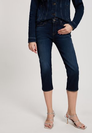 Dunkelblaue Capri-Jeans mit einer figurbetonten Passform, fünf Taschen und einem umgeschlagenen Saum. Kombiniert mit silbernen, geschnürten High-Heels.