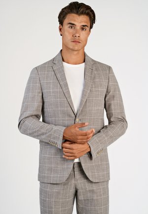 Mies pukeutunut vaaleanharmaaseen ruutukuosiseen pukuun ja valkoiseen paitaan, seisoo käsivarret ristissä vyötäröllä yksinkertaisen taustan edessä.