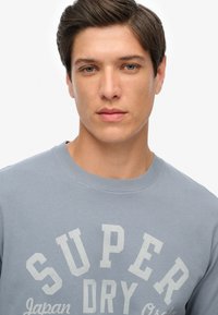 Sudadera azul claro con cuello redondo, que presenta un gran logo impreso en blanco que dice "SUPER DRY" y un texto más pequeño debajo.