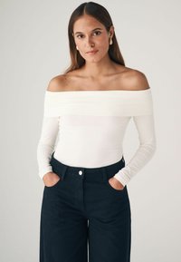 Fehér, off-the-shoulder, hosszú ujjú felső, testhez simuló fazonnal, magas derekú sötét farmernadrággal viselve, ami zsebekkel és gombos zárással rendelkezik.