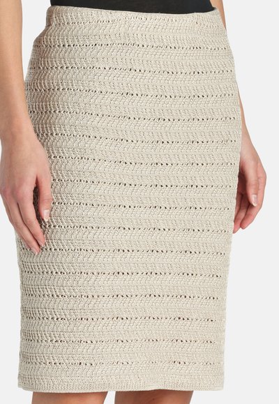 Jupe beige au crochet longueur genou avec des motifs en zigzag horizontaux portée par une personne les mains détendues le long du corps.