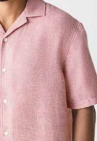 Camisa de manga corta en tejido ligero de color rojo claro con un acabado texturizado, que presenta un cuello de punta y botones blancos en la parte frontal.