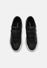 Black leather sneakers with three velcro straps, white rubber soles, rounded shape, and smooth texture. Branding visible on insoles.