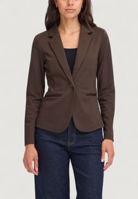Blazer brown aderente con rever a punta, chiusura a un bottone e due tasche frontali, abbinato a pantaloni in denim scuro. Texture liscia.