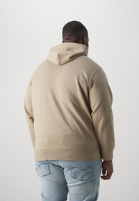 Beige Kapuzenpullover mit lockerer Passform. Weicher Stoff, gerippter Saum und Bündchen. Kombiniert mit hellblauen Jeans mit Rückentaschenstickerei.