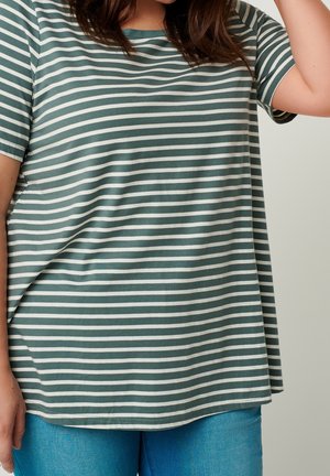 Henkilö, jolla on yllään vihreä-valkoraidallinen, löysä vaakasuuntainen t-paita ja siniset farkut, toinen käsi kohotettuna pään lähellä.