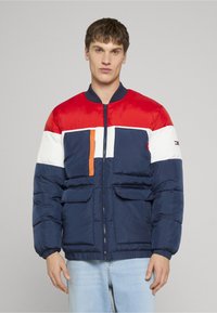 Námornícky modrá prešívaná bunda s červenými, bielymi a oranžovými akcentmi. Má zapínanie na zips, dve predné vrecká a rebrované manžety. Ľahká, hladká na dotyk.
