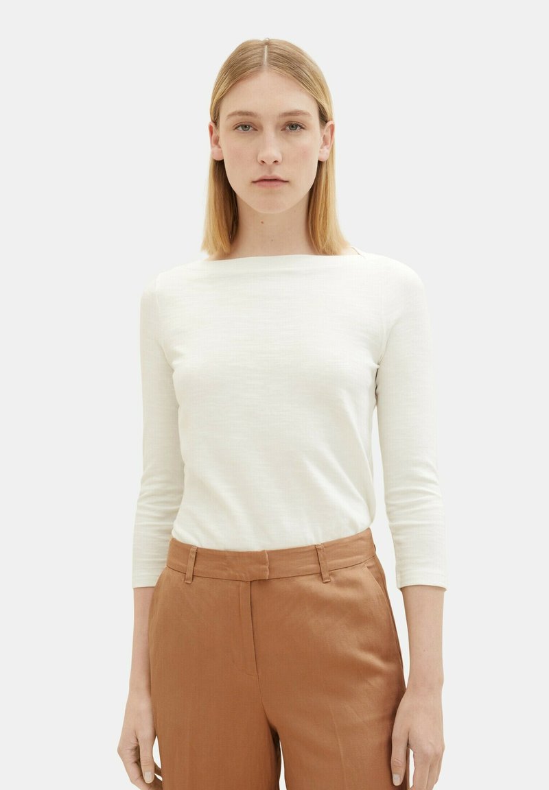 Haut long à manches blanches avec un col bateau, associé à un pantalon taille haute marron. Le tissu semble texturé et léger.