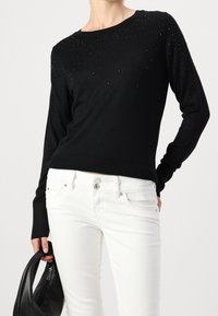 Pull noir à manches longues avec des embellissements texturés sur les épaules, associé à un jean skinny blanc et un sac noir. Tissu en maille lisse.