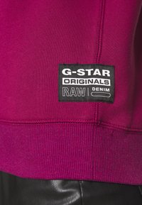 Fuchsia sweatshirt med ribbad fåll och ärmslut, har en svart tygetikett med vit text: "G-STAR ORIGINALS RAW DENIM."
