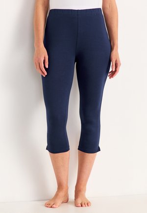 Kvinde iført mørkeblå leggings til midt på læggen med elastisk linning, står barbeint mod en hvid væg.