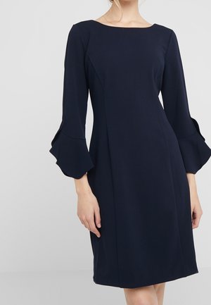 Frau trägt ein marineblaues knielanges Kleid mit dreiviertel Ärmeln, die mit Rüschen an den Bündchen versehen sind, und einem U-Boot-Ausschnitt.