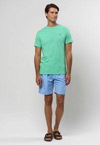 Hele rohelise puuvillase T-särgiga, millel on väike sinine logo, kombineeritud helesiniste lühikesed püksid. Sandaalid tumedate pruuni rihmadega.