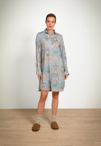 Vrouw in een lange mouw, met een patroon gekleed knielang jurkje, met beige sokken en bruine pantoffels, staand op een houten vloer tegen een witte muur.