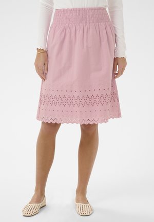 Vrouw draagt een roze rok tot op de knie met bloemrijke oogjespatronen en een geschulpte zoom, gecombineerd met een wit topje met lange mouwen en crème gekleurde geweven platte schoenen.