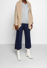 Trench coat beige aperto sopra una maglietta a righe bianche e blu navy, abbinato a pantaloni in denim scuro a gamba larga e stivaletti bianchi.