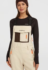 Beige overall met een bruine borstzak en oranje ritsaccenten. Verstelbare bandjes en zijzakken vergroten de functionaliteit. Zwarte lange mouwen top eronder.