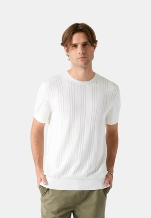 Uomo con capelli castani indossa una maglietta bianca a maniche corte lavorata a maglia con righe verticali e pantaloni verde oliva, in piedi con le mani in tasca.