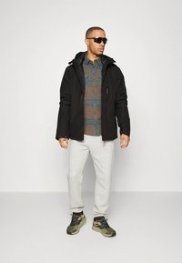 Zwarte jas met ritssluiting en capuchon, gedragen over een bruin-blauw geruit overhemd. Grijze joggingbroek en sneakers met camouflagemotief. Zwarte zonnebril.