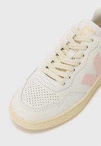 White leather sneaker with perforated toe, cream laces, and light pink accents. Features a textured sole and stitched detailing.