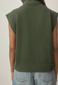 Hladký svetřík bez rukávů ve olivově zelené barvě, pletený rolák, nošený s lehkými džíny, zobrazený zezadu na osobě.