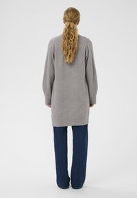 Maglione grigio a maglia con una vestibilità comoda, spalle scese e polsi a coste, abbinato a jeans larghi blu scuro. Sfondo semplice.