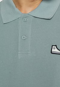 Polo shirt bleu clair avec un tissu texturé, deux boutons et un logo de basket noir et blanc sur la poitrine à gauche.