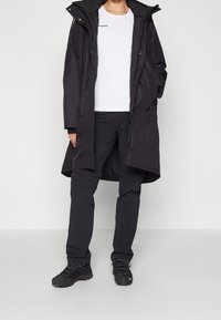 Manteau isolant noir avec capuche, associé à une chemise blanche et un pantalon noir. Dispose d'une fermeture éclair et d'un tissu durable, adapté pour une utilisation en extérieur.