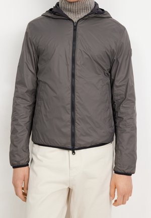 Homme portant une veste à capuche grise zippée par-dessus un col roulé beige et un pantalon de couleur claire, debout les bras détendus le long du corps.