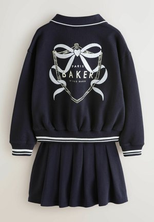 Marineblå kjole med en bomberinspireret overdel og en plisseret nederdel. Har hvide broderede sløjfer og striber ved ærmekanter og taljebånd.