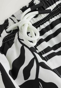 Primo piano di pantaloncini da bagno in tessuto a fantasia bianca e nera con coulisse bianca annodata e estremità in plastica nera con la scritta "NEXT".