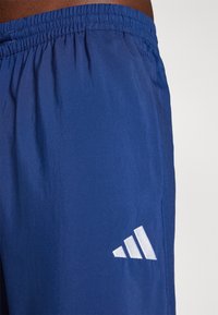 Modre atletske kratke hlače iz lahkega materiala, z elastičnim pasom in belim logotipom s tremi črtami na strani.