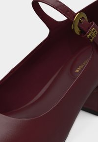 Κλειστό παπούτσι με τακούνι σε μπορντό δέρμα, με λουράκι στον αστράγαλο και λεπτομέρεια με χρυσή αγκράφα, με λεία επιφάνεια και τακούνι τύπου block.