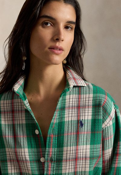 Femme aux longs cheveux foncés portant des boucles d'oreilles beige en perles et cristal et une chemise à carreaux verte, blanche et rouge avec col et boutons.