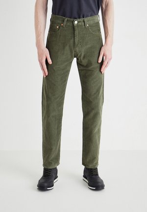 Mann trägt olivgrüne Cordhose, schwarze Schuhe und ein schwarzes Hemd, steht mit entspannten Armen seitlich am Körper vor weißem Hintergrund.