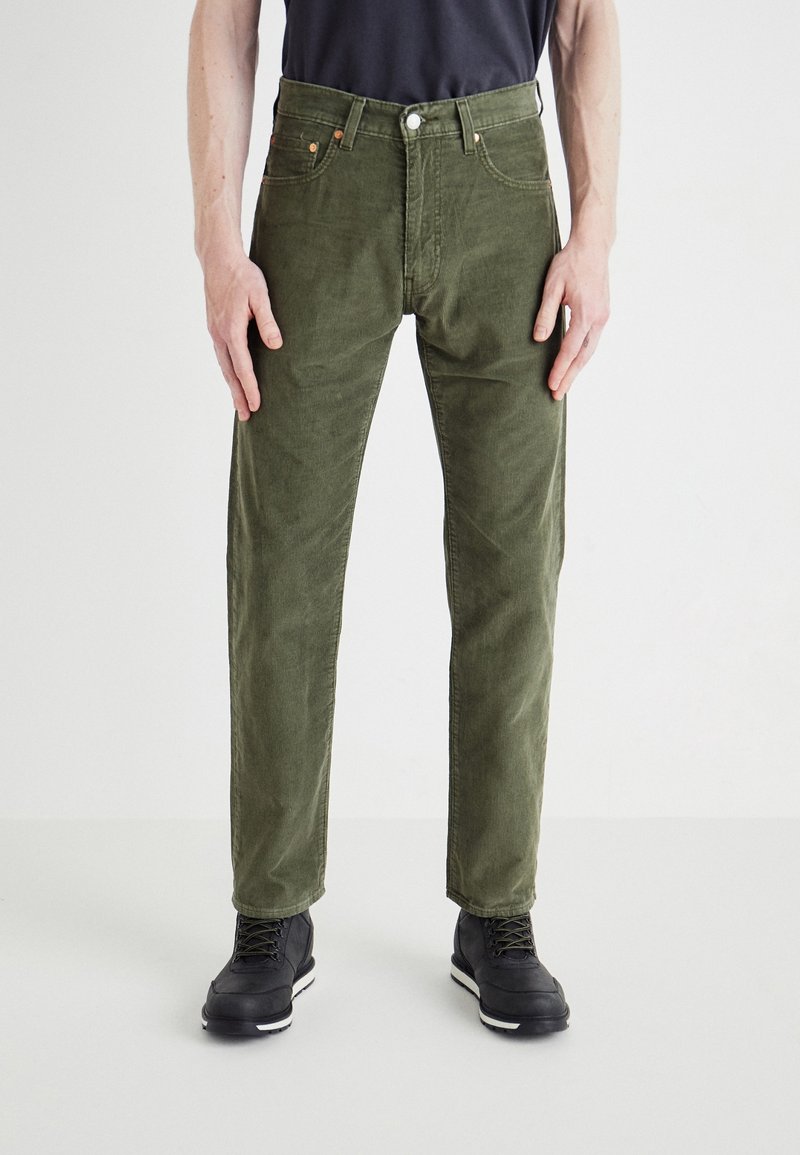 Homme portant un pantalon en velours côtelé vert olive, des chaussures noires et une chemise noire, debout avec les bras détendus le long du corps sur fond blanc.