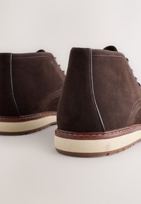 Braune hochgeschnittene Wildlederschuhe mit einer hellfarbenen Gummisohle, kontrastierenden Nähten und einer strukturierten Oberfläche an Ferse und Seiten.