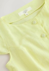 Blouse en lin jaune pâle avec encolure ronde, deux boutons recouverts de tissu, manches courtes et étiquette de marque visible à l'intérieur du col.