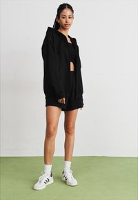 Juodas zip-up džemperis su priekinėmis kišenėmis, uždėtas ant juodo trumpomis rankovėmis topuko ir šortų. Balti sportbačiai su tamsiais akcentais. Laidūs kontūrai, švelni tekstūra.