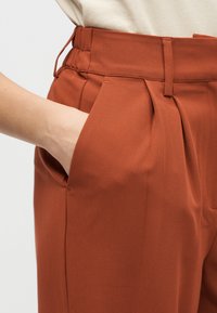 Παντελόνι σε χρώμα σκουριάς με ψηλή μέση, διαθέτει πιέτες, πλαϊνή τσέπη και λείο, υφασμάτινο ύφασμα με υφή.