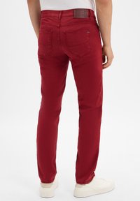 Homme portant un pantalon rouge slim et des baskets blanches, se tenant de dos contre un fond blanc.