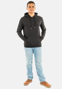 Haut à capuche noir avec cordons de serrage, poche frontale et détail de logo, associé à un jean bleu clair et des chaussures marron, sur fond uni.