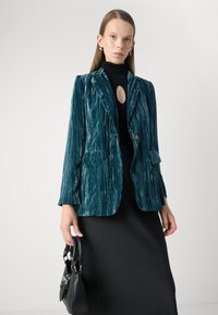 Teal velvet blazer with vertical stripes, notched lapels, and two front pockets. Paired with a black top and skirt, held with a black handbag.