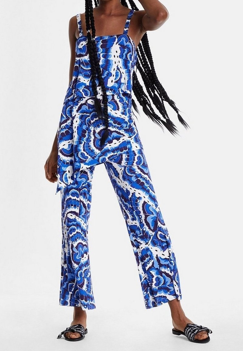 Blå och vit mönstrad jumpsuit med vida ben och tunna axelband, tillverkad av ett glatt, lätt material. Bärs med svarta platta sandaler.