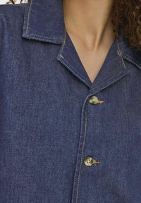 Camicia di jeans blu scuro con colletto, caratterizzata da cuciture gialle e due bottoni rotondi sul davanti. Vista ravvicinata della texture e dei dettagli.