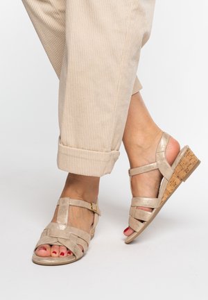 Beige wedge sandals with ankle straps worn with cuffed beige corduroy pants on crossed feet.